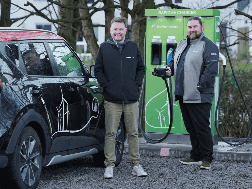 Electric vehicle parked at a rapid EV charging station with two people standing beside the charger, illustrating public EV charging infrastructure and support for electric vehicle adoption.