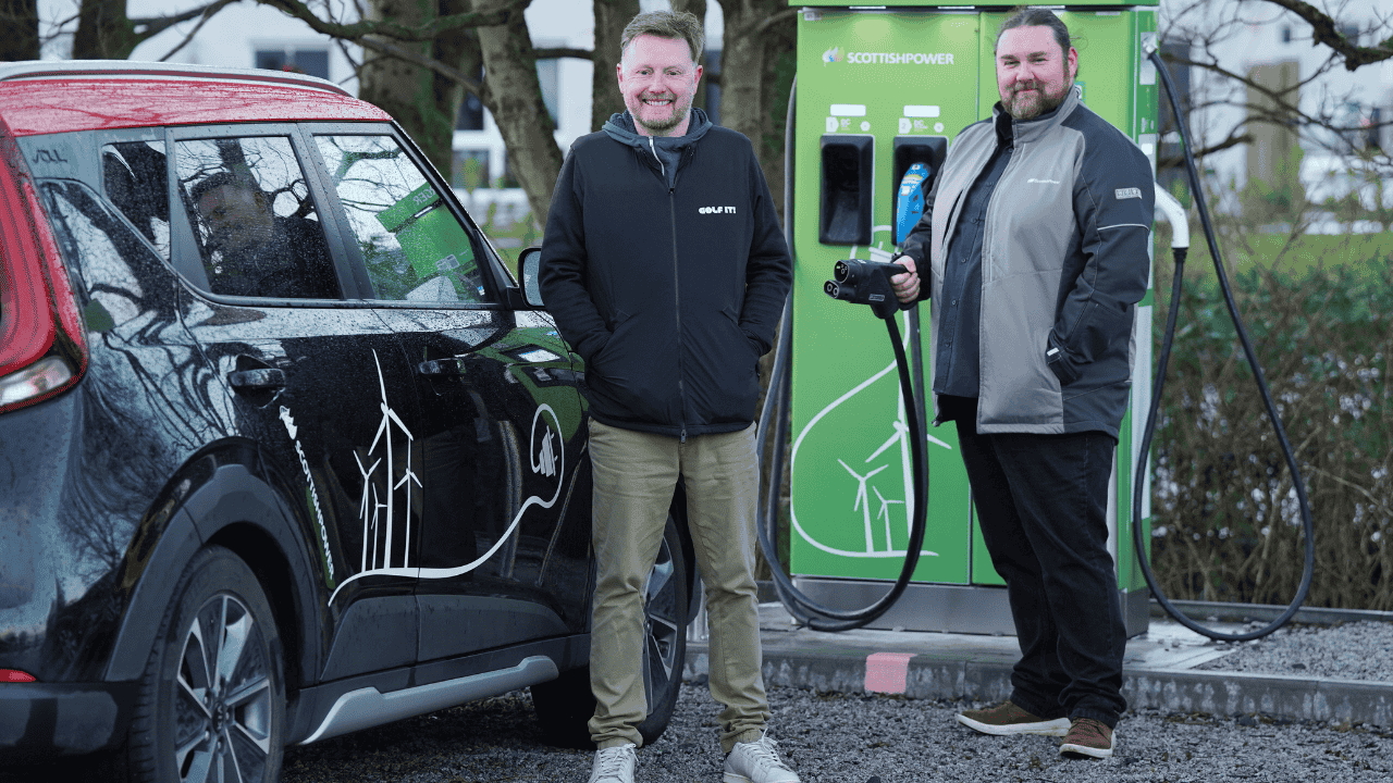 Electric vehicle parked at a rapid EV charging station with two people standing beside the charger, illustrating public EV charging infrastructure and support for electric vehicle adoption.