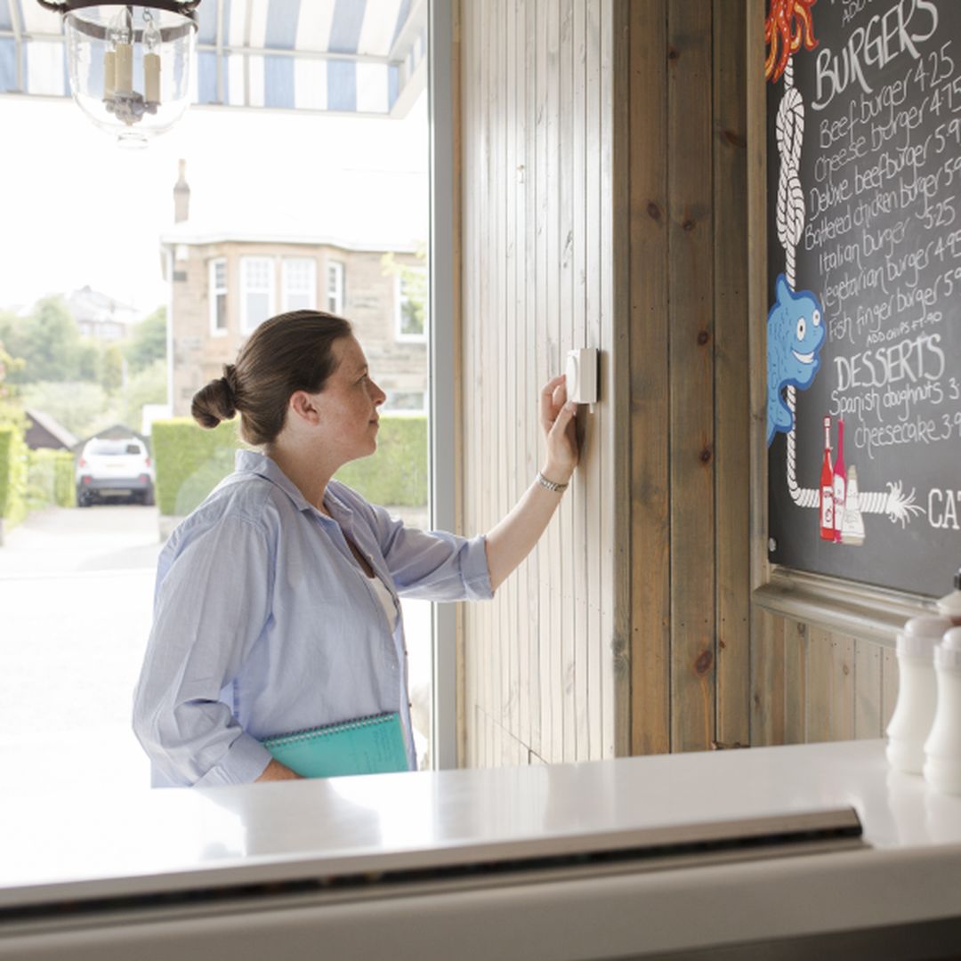 A person stands inside a restaurant, looking at a wall-mounted menu board. The counter in front has condiments like salt, pepper, and sauce bottles.