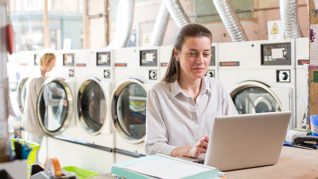 Person using a laptop in a launderette with washing machines in the background, managing everyday tasks