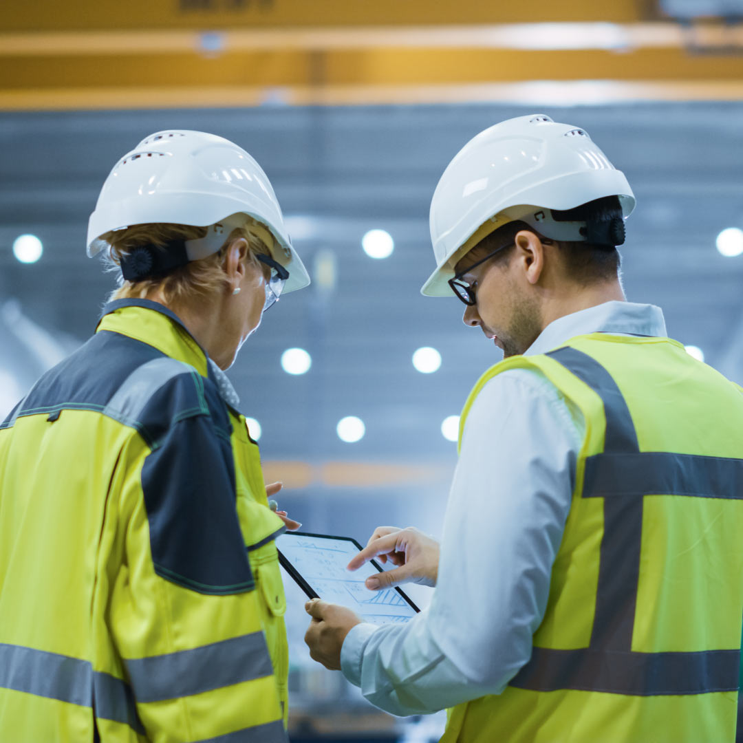 Workers in safety gear reviewing plans on a tablet at an industrial site.