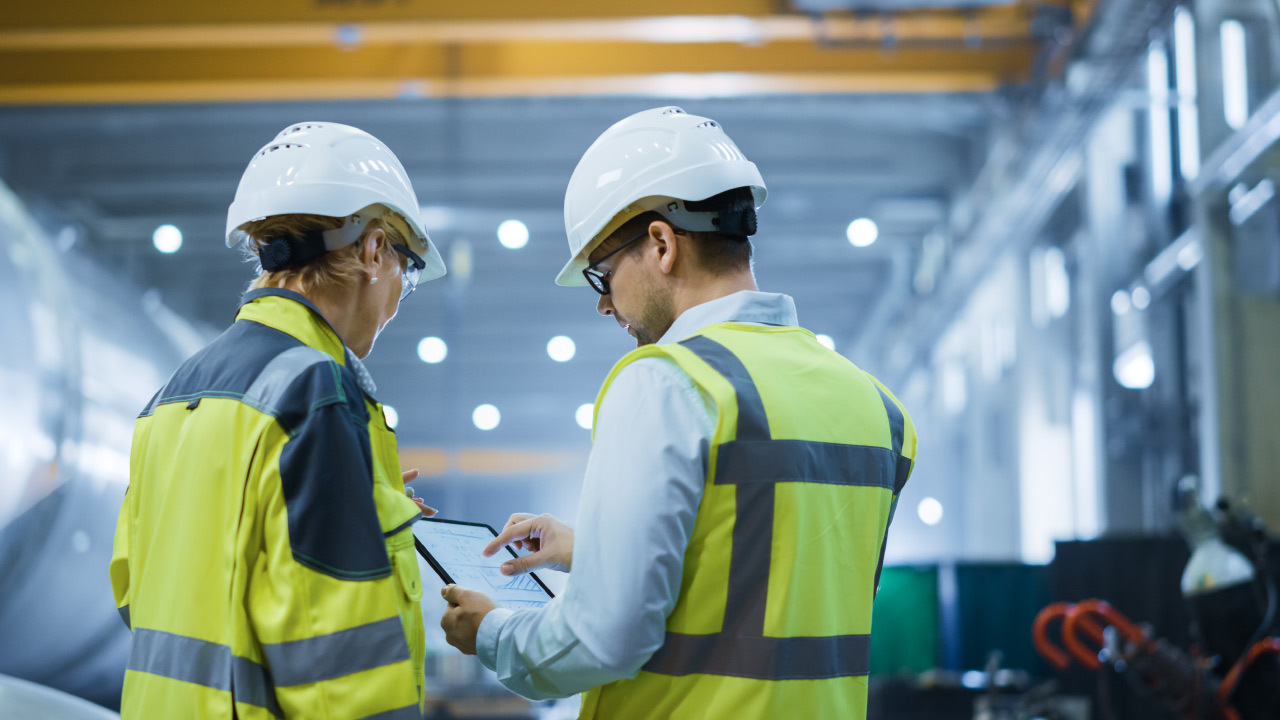 Workers in safety gear reviewing plans on a tablet at an industrial site.