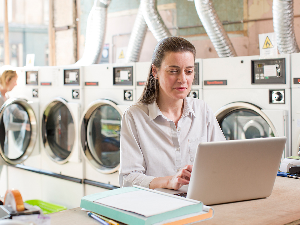 Person using a laptop in a launderette with washing machines in the background, managing everyday tasks