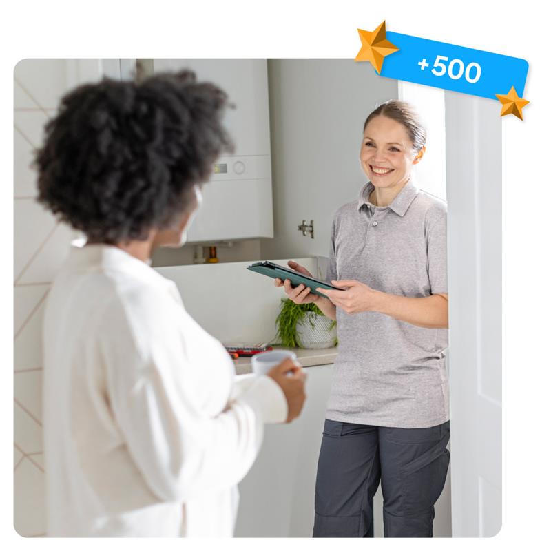 Home energy assessment taking place indoors, with a person holding a tablet and speaking to a homeowner near a wall-mounted boiler, with a blue ‘+500’ badge in the top corner.