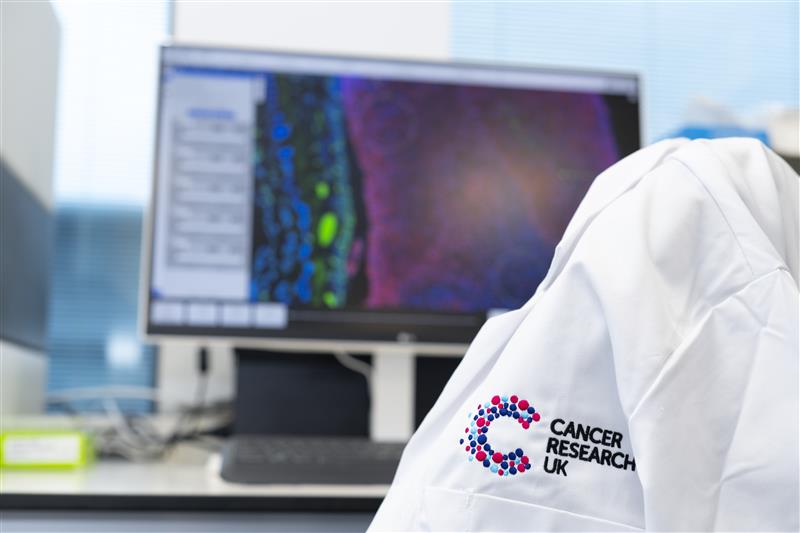 A white laboratory coat with the Cancer Research UK logo draped over a chair in front of a computer monitor.