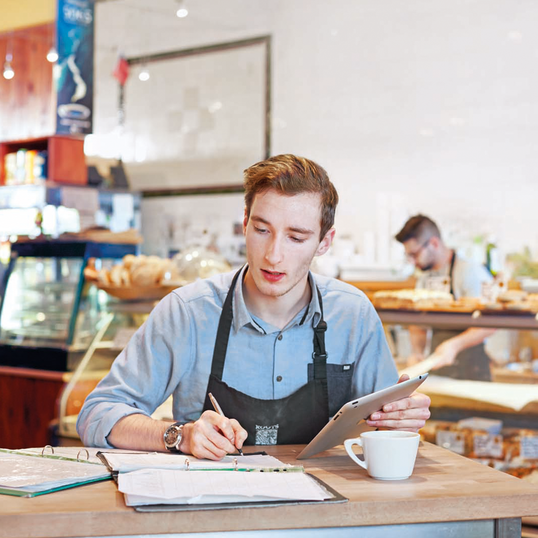 Bakery staff using tablet and writing at table with documents and coffee, shelves of bread and pastries in background