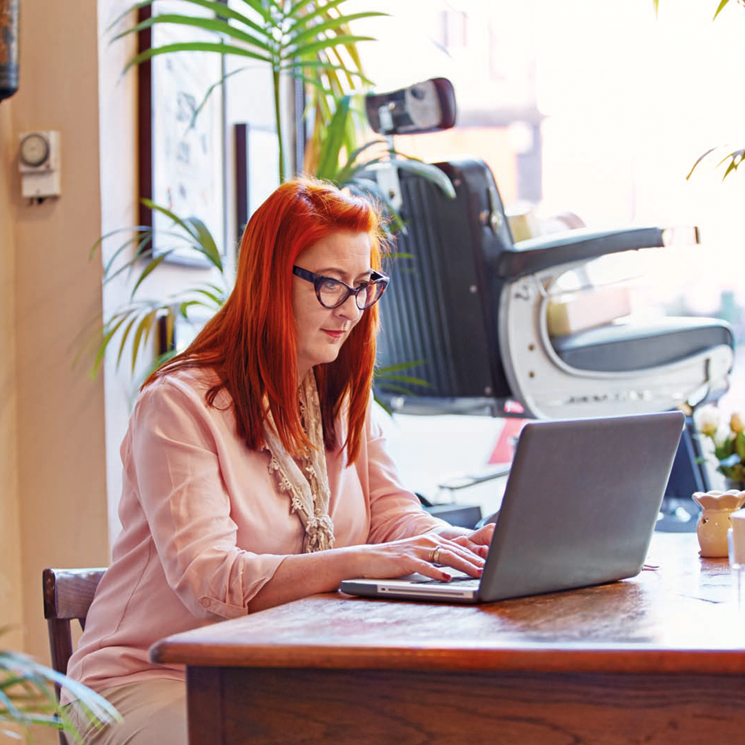 Woman with red hair and glasses working on a laptop at a wooden desk in a cozy, well-lit room