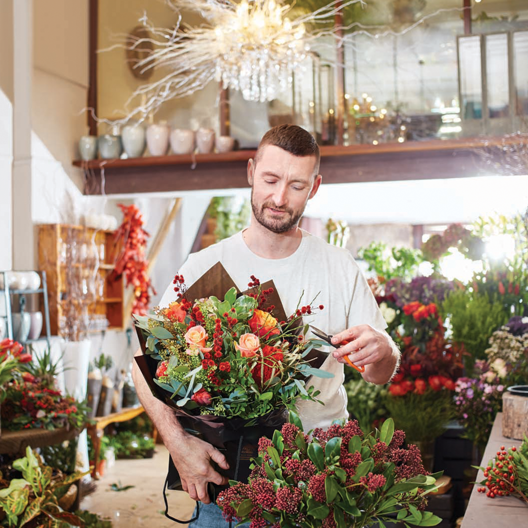 Male florist holding bouquet in flower shop, surrounded by floral arrangements and plants