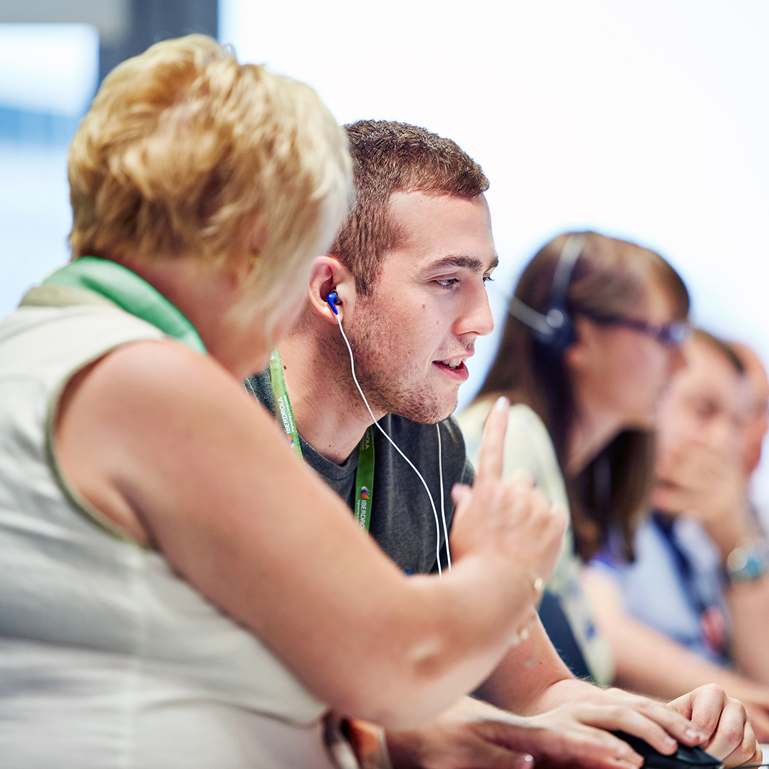 A group of support agents seated at desks, wearing headsets and lanyards. One agent in the foreground is speaking and gesturing toward a colleague while the team handles customer calls.