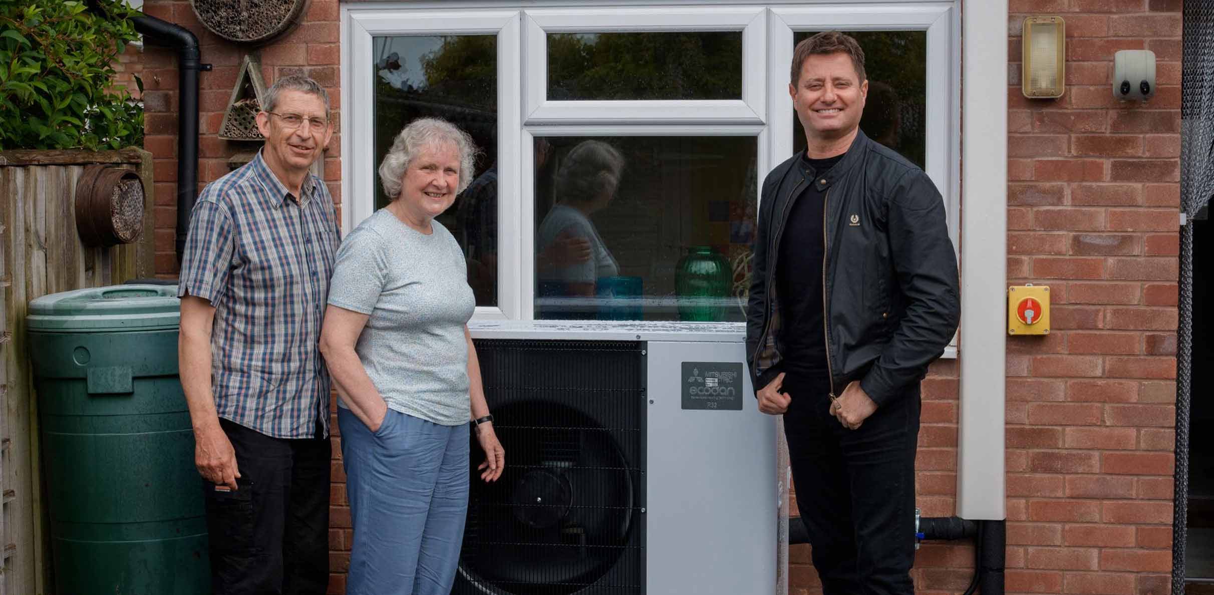 Man and woman couple standing in front of a heat pump in their garden with George Clarke