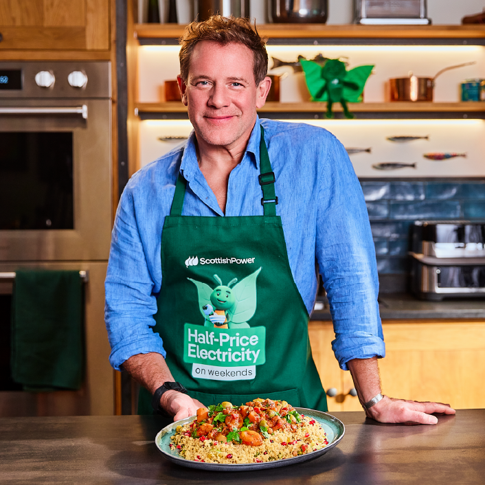 TV chef Matt Tebbutt in a Bruce apron holding a plate of vegetable tagine and fruity couscous in a kitchen.