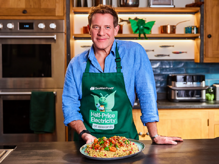 TV chef Matt Tebbutt in a Bruce apron holding a plate of vegetable tagine and fruity couscous in a kitchen.