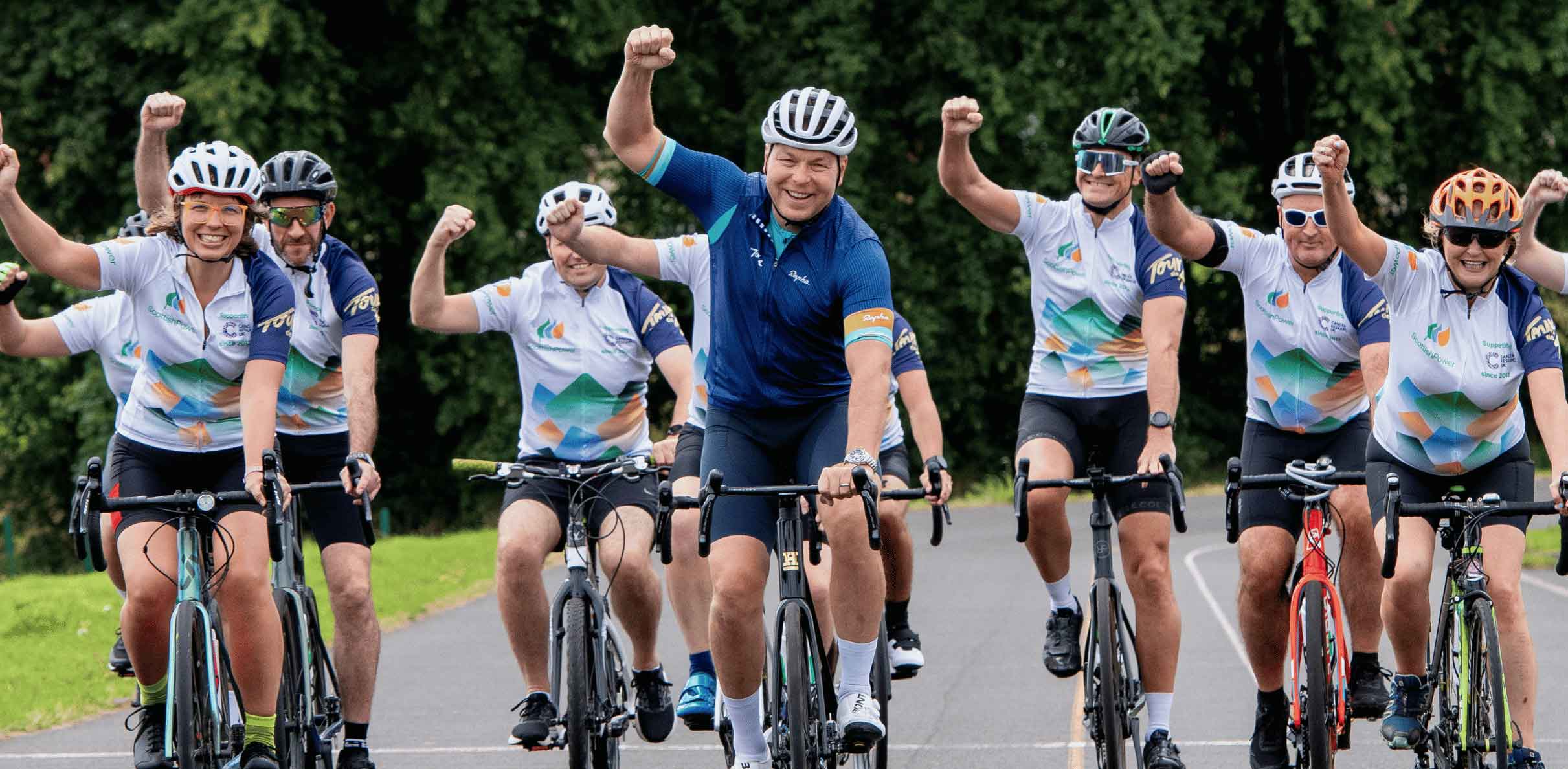 Group of cyclists riding together on a country road during a charity cycling event, wearing helmets and matching cycling jerseys while raising their arms as they pedal through a tree-lined route.