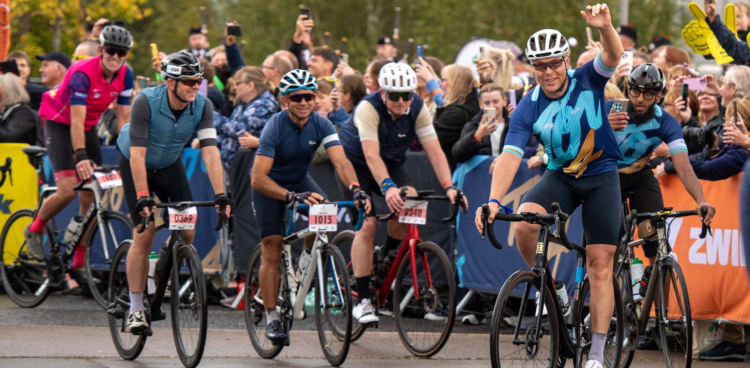 Cyclists riding past cheering spectators during a large organised road cycling event, with participants wearing helmets and numbered race bibs as crowds line the route taking photos and showing support.